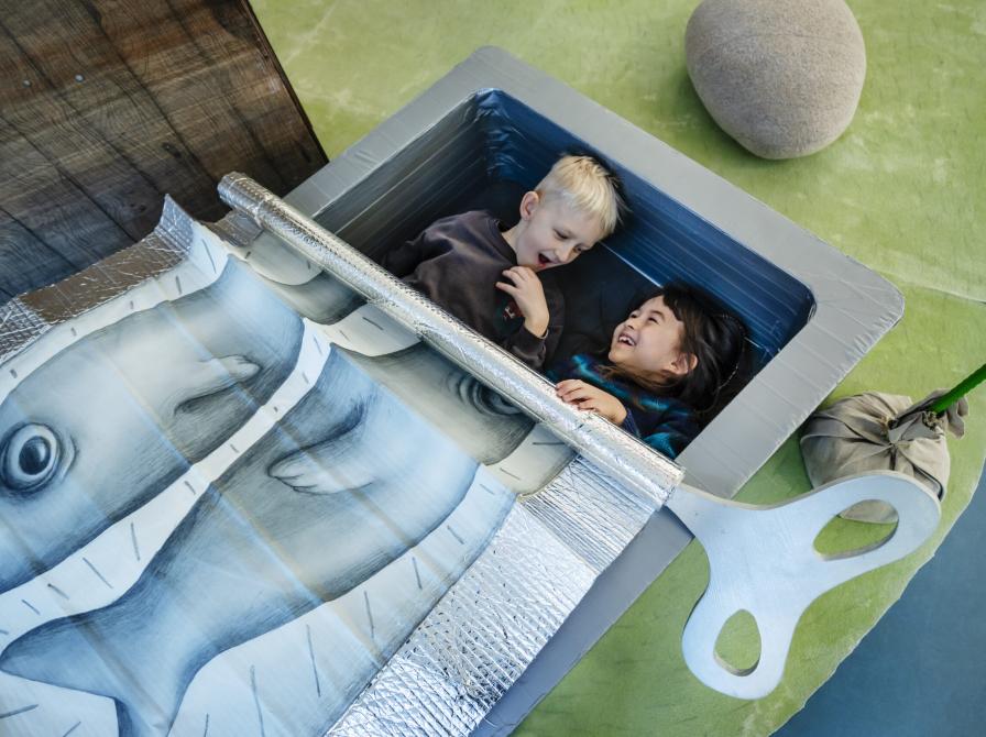 Foto fra installation: To børn ligger i en kæmpe sardindåse og leger sardiner.
